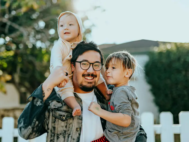 Pai sorrindo com os dois filhos no colo, em frente de casa